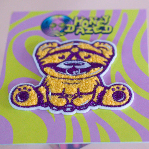 Patch featuring a cute bear in yellow and purple with a playful expression, wearing a BDSM harness and a ball gag. The design is fun and whimsical, combining bold colors with a cheeky style.