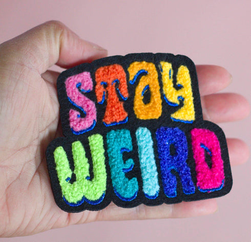Colorful patch with the phrase 'Stay Weird' in vibrant, multicolored, textured letters, set against a black background. The design is playful and bold, celebrating uniqueness and individuality.
