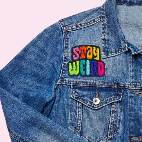 Colorful patch with the phrase 'Stay Weird' in vibrant, multicolored, textured letters, set against a black background. The design is playful and bold, celebrating uniqueness and individuality.