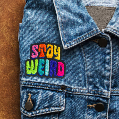 Colorful patch with the phrase 'Stay Weird' in vibrant, multicolored, textured letters, set against a black background. The design is playful and bold, celebrating uniqueness and individuality.