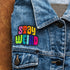 Colorful patch with the phrase 'Stay Weird' in vibrant, multicolored, textured letters, set against a black background. The design is playful and bold, celebrating uniqueness and individuality.