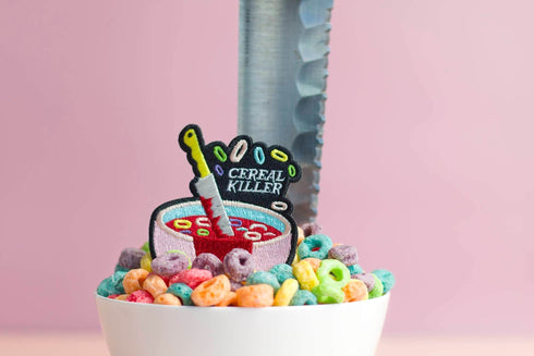Patch featuring a bowl of cereal with a dripping knife and the text 'Cereal Killer,' set against a light pink background. The design combines playful cereal elements with a humorous, spooky twist.