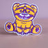 Patch featuring a cute bear in yellow and purple with a playful expression, wearing a BDSM harness and a ball gag. The design is fun and whimsical, combining bold colors with a cheeky style.