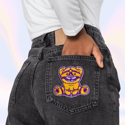 Patch featuring a cute bear in yellow and purple with a playful expression, wearing a BDSM harness and a ball gag. The design is fun and whimsical, combining bold colors with a cheeky style.