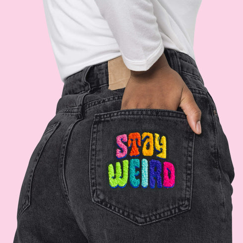 Colorful patch with the phrase 'Stay Weird' in vibrant, multicolored, textured letters, set against a black background. The design is playful and bold, celebrating uniqueness and individuality.