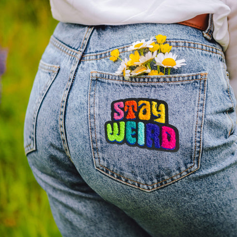 Colorful patch with the phrase 'Stay Weird' in vibrant, multicolored, textured letters, set against a black background. The design is playful and bold, celebrating uniqueness and individuality.