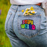 Colorful patch with the phrase 'Stay Weird' in vibrant, multicolored, textured letters, set against a black background. The design is playful and bold, celebrating uniqueness and individuality.