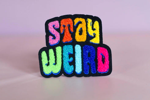 Colorful patch with the phrase 'Stay Weird' in vibrant, multicolored, textured letters, set against a black background. The design is playful and bold, celebrating uniqueness and individuality.