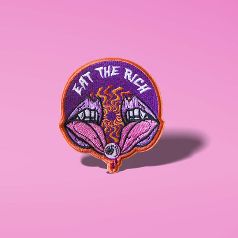 Patch featuring the text 'Eat The Rich' above a design of two open mouths with fangs and a tongue holding an eyeball, set against a purple background with an orange border. The design is bold and edgy.