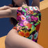 A colorful one-piece swimsuit features a vibrant, quirky pattern with cherries, cartoon eyes, and playful designs on a dark background.