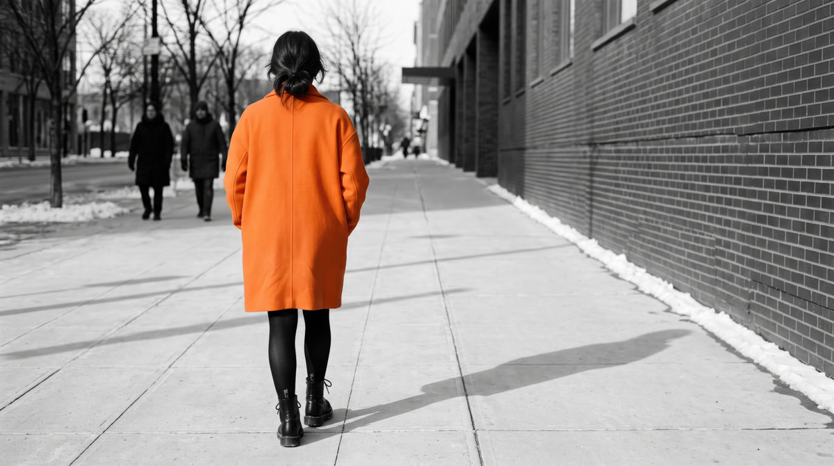 Woman in a bright tangerine wool coat and black combat boots walking down a gray winter city sidewalk