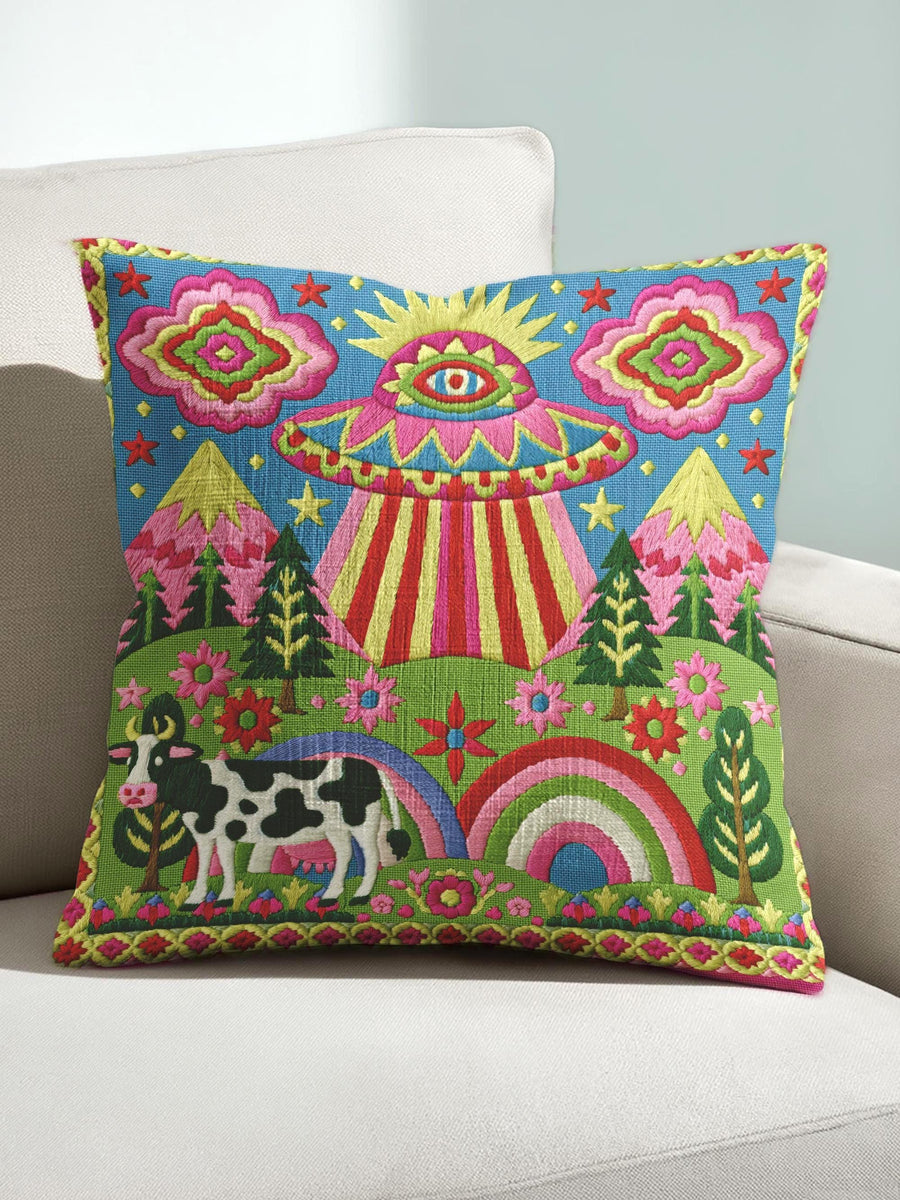 Description of a square throw pillow with a vibrant maximalist alien design. A central UFO with a large all-seeing eye hovers above a landscape with two rainbows, pink mountains, and green pine trees. A black and white spotted cow stands in the foreground. The design is a high-contrast palette of sky blue, magenta, hot pink, and meadow green. The pillow has a faux-embroidery 2D print finish on linen-feel fabric, framed by a yellow and pink scalloped floral border.