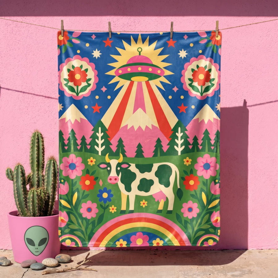 A rectangular velveteen minky blanket with a bright, maximalist design. The top half features a vibrant blue sky with a pink, green, and yellow UFO beaming rays down over hot pink snow-capped mountains. The bottom half shows a grassy green field with a white cow covered in dark spots, surrounded by large, colorful red, pink, and blue retro flowers, pine trees, and a rainbow arch at the very bottom. The reverse side is a fluffy off-white sherpa.