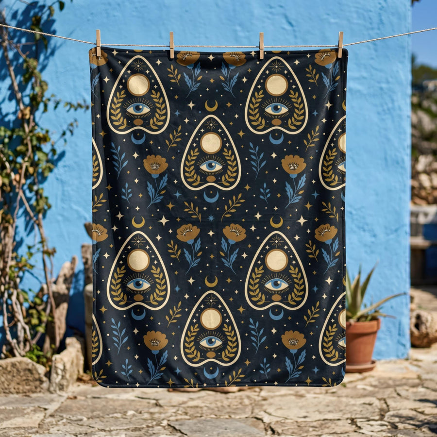 A rectangular velveteen minky blanket featuring a repeating dark academia pattern on a deep black background. The design includes gold and cream Ouija planchettes, each containing a blue all-seeing eye, a cream circle, and a light blue crescent moon framed by botanical leaves. Scattered between the planchettes are gold flowers with light blue stems, additional crescent moons, and small four-point stars. The reverse side of the blanket is made of a fluffy, textured off-white sherpa fleece.
