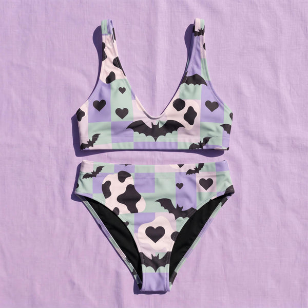 A two piece recycled high waisted swimsuit with a pastel goth checkered print in light lavender purple, pale mint green, and white squares with black cow print spots. The design features small black hearts and silhouetted bats with outspread wings printed throughout. The athletic style crop top has a deep scoop neck and matching thick straps, while the double layered bottoms have a supportive high waistband and cheeky cut with a solid black inner lining.