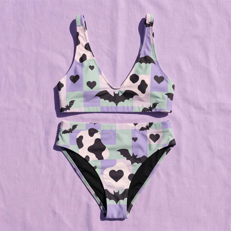 A two piece recycled high waisted swimsuit with a pastel goth checkered print in light lavender purple, pale mint green, and white squares with black cow print spots. The design features small black hearts and silhouetted bats with outspread wings printed throughout. The athletic style crop top has a deep scoop neck and matching thick straps, while the double layered bottoms have a supportive high waistband and cheeky cut with a solid black inner lining.