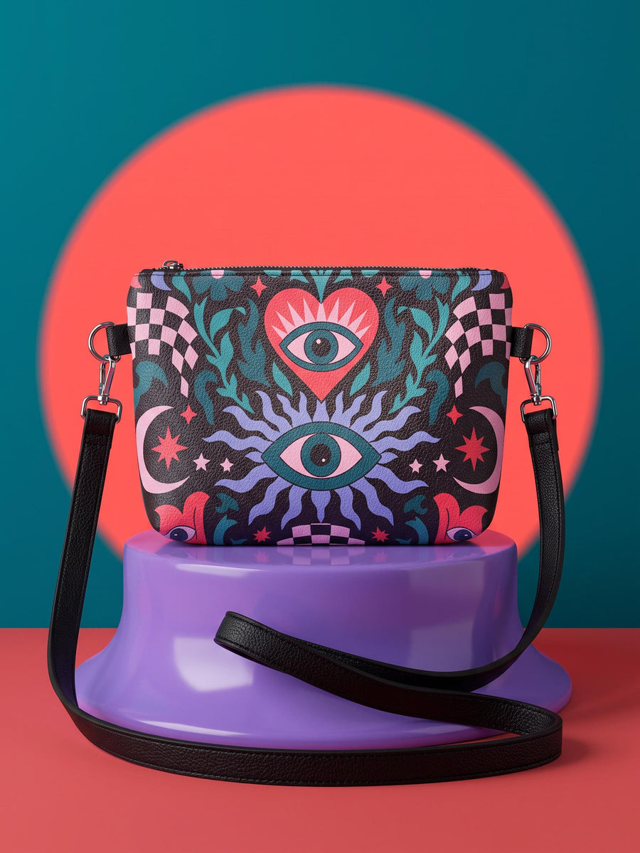 Small vegan leather crossbody purse with a real pebble grain texture and adjustable black strap. The flat printed artwork features a central blue sunburst evil eye motif below a coral pink sacred heart eye, surrounded by pink crescent moons, multicolored stars, flowing teal botanical vines, and wavy lavender and black checkerboard distortion banners. Set against a dark background with silver metal hardware including a top zipper, zipper pull, and strap clips. Measures 8.5 by 7 by 2 inches.
