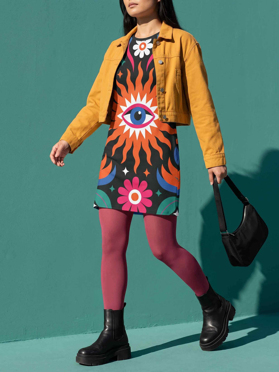 A short-sleeve, above-the-knee t-shirt dress with a straight, structured fit. The dress has a true black background featuring a bold, vibrant graphic pattern. The center displays a large blue and white evil eye surrounded by a bright orange wavy sunburst with a white jagged border. Above and below the eye are bright pink daisy flowers with orange centers, scattered white four-petal stars, and wavy blue and teal green leaf shapes. The fabric is thick, smooth, and holds its clean shape.