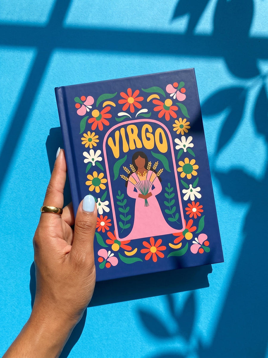 Royal blue hardcover journal featuring a retro folk art design. A center pink arch frames the word VIRGO in golden groovy rounded lettering. Below sits an illustrated maiden with brown skin and dark hair, wearing a pink dress and holding a bundle of golden wheat. A dense floral border wraps the cover with daisies and blooms in orange, marigold yellow, cream white, and pink, accented by green leaves and stems. Matte finish cover with a blue spine reading Virgo. Lined pages inside.