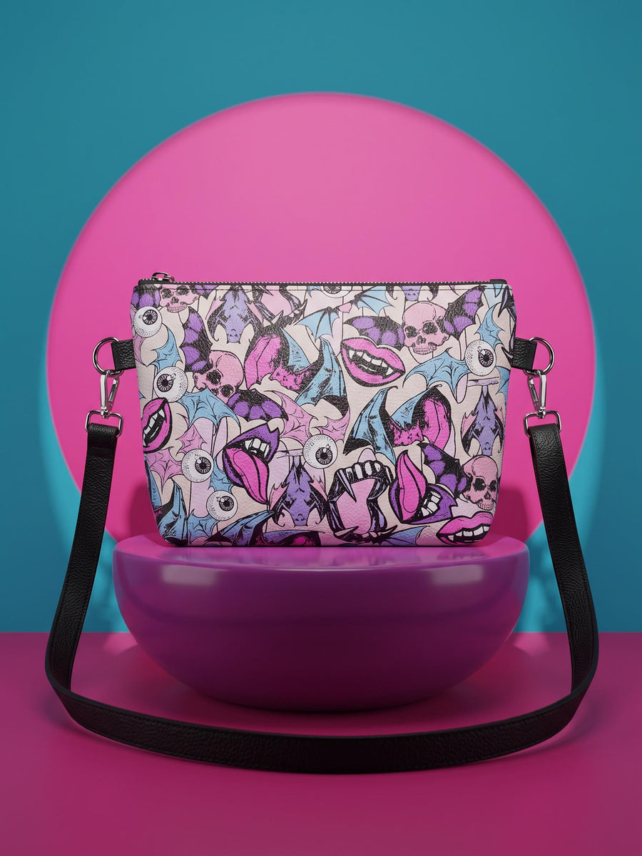 Small vegan leather crossbody purse with a real pebble grain texture and adjustable black strap. The flat printed artwork features a scattered all over pattern of pink skulls, purple and pastel blue winged bats, pink fanged mouths, and loose eyeballs set against a cream colored background. The bag is shown with black and silver hardware including a black top zipper, black zipper pull, black strap tabs, and silver metal rings and clips. The compact rectangular bag measures 8.5 by 7 by 2 inches.