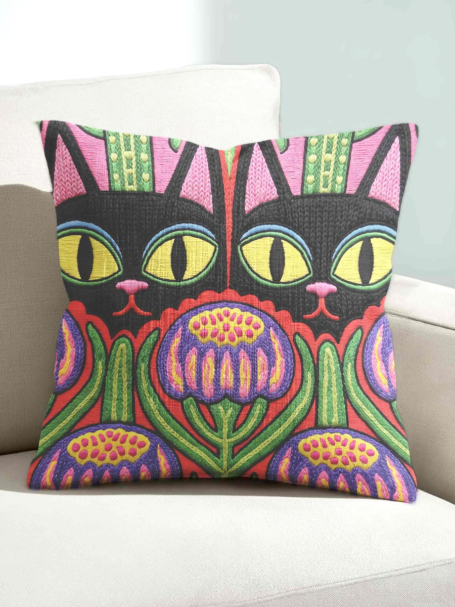 A square decorative pillow cover featuring a vibrant, symmetrical flat printed design that mimics the look of textured embroidery. Two black cats with large yellow eyes and pink noses face forward against a bright red background. Below the cats are large purple and pink flowers with green stems. Above the cats are green cactus shapes. The artwork uses bold, colorful outlines to create an optical illusion of stitching, though the fabric itself is smooth.