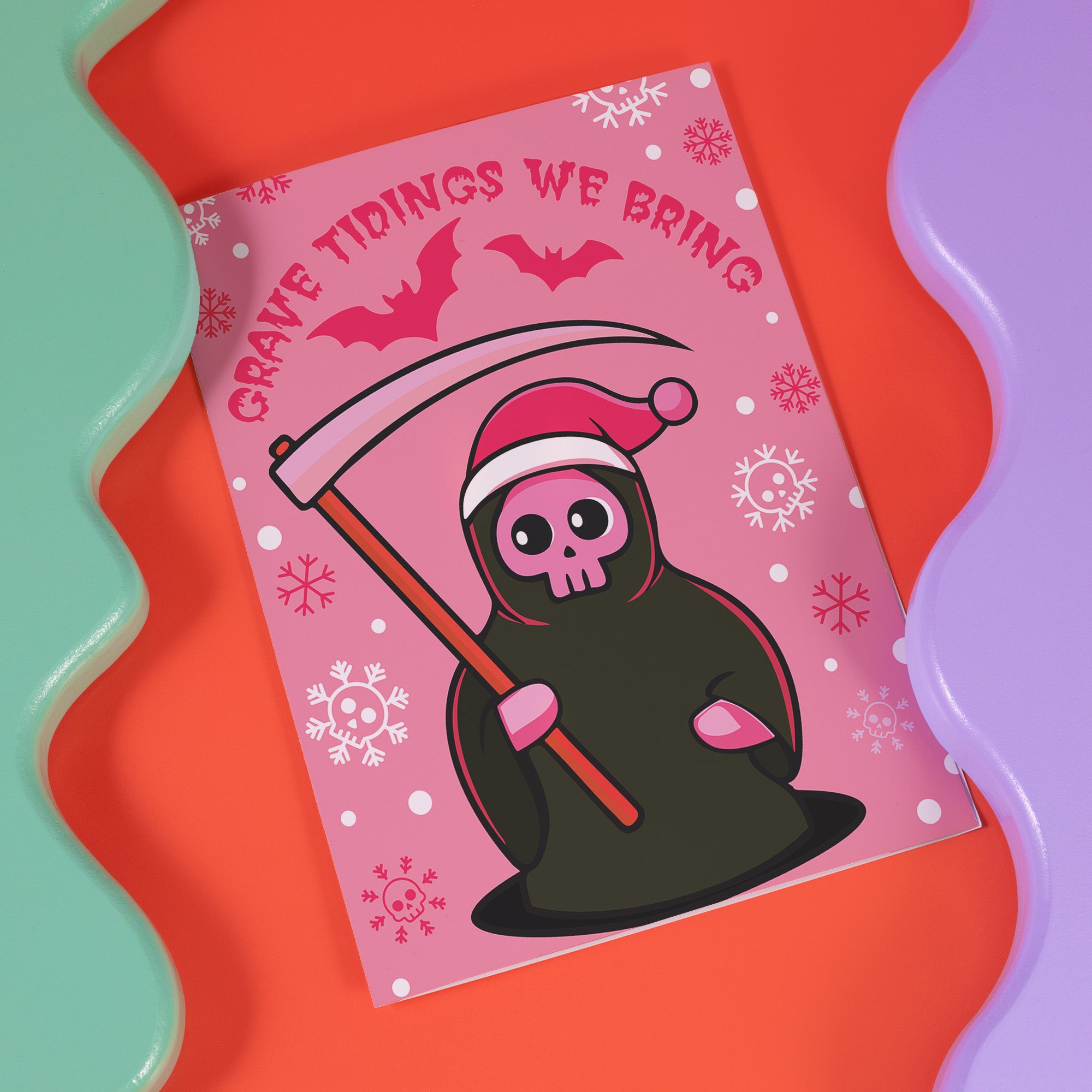 Quirky Christmas card featuring grim reaper in Santa hat. Pink background with bats, snowflakes, skull motifs. Text reads 'Grave Tidings We Bring'. Humorous blend of holiday cheer and gothic aesthetic. Perfect for alternative Christmas greetings.