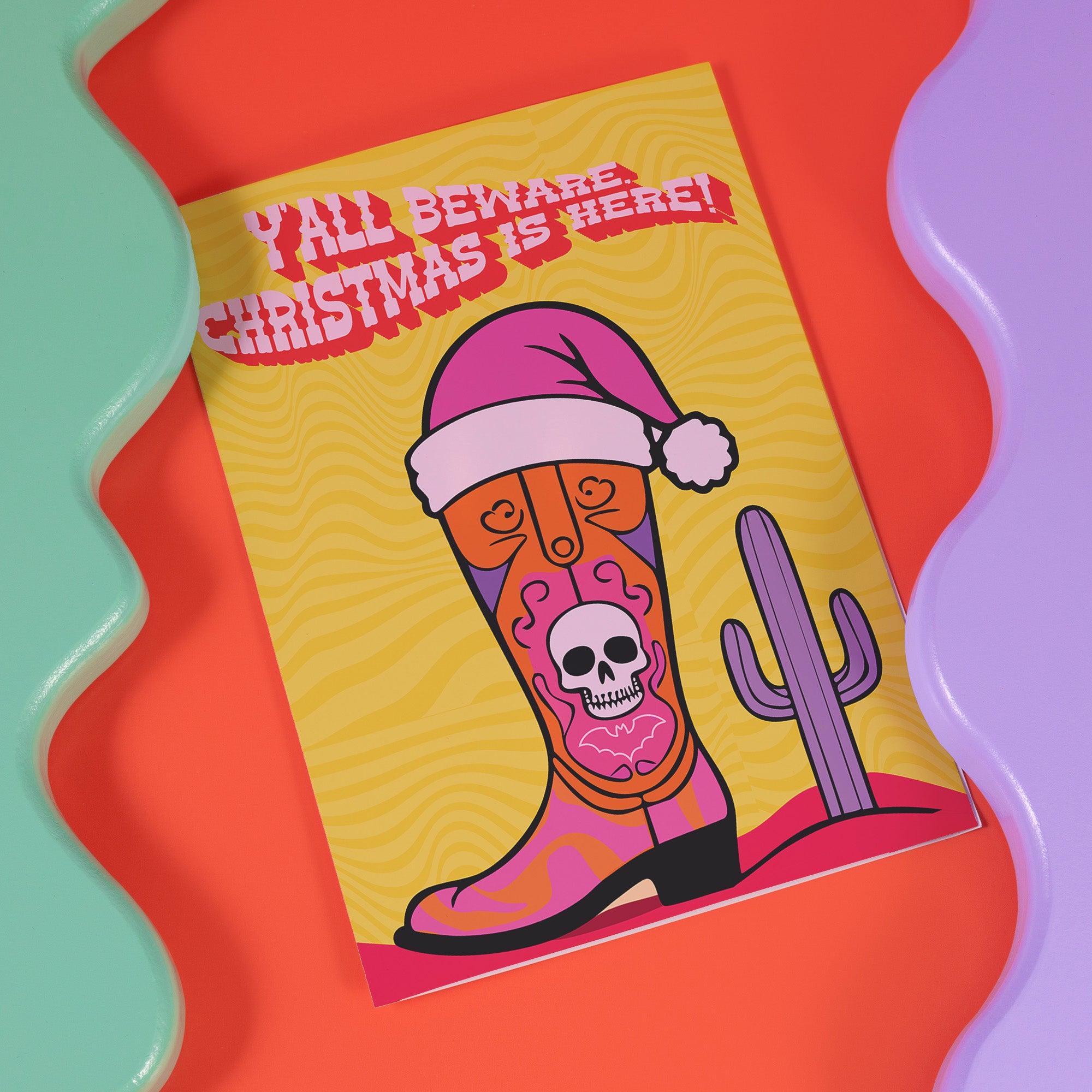 Colorful Christmas card with a yellow background. Features a stylized cowboy boot decorated with a skull, hearts, and swirls in pink, orange, and purple. The boot wears a pink Santa hat. A green cactus stands beside the boot. Top text reads 'Y'ALL BEWARE, CHRISTMAS IS HERE!' in red, wavy letters. Card has a purple border. Inside message: 'May Your days be merry, bright and full of southern delight.' The design blends Christmas, Western, and Day of the Dead motifs for a unique, playful holiday greeting