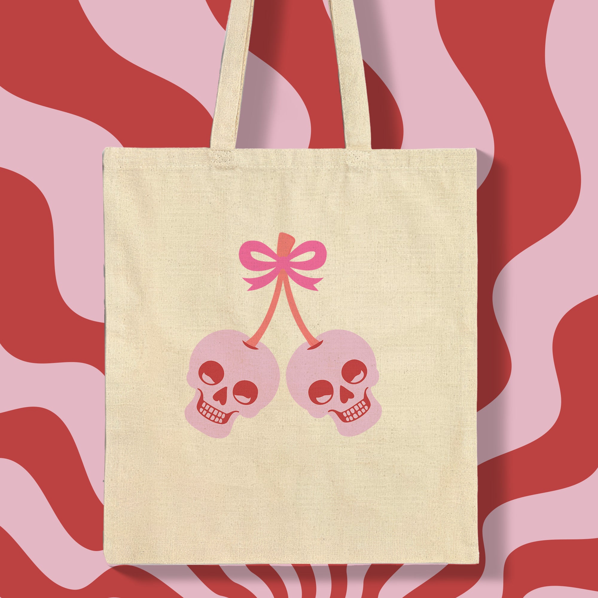 Canvas tote bag featuring pink skull cherries design. Two pink skulls connected by red stem and bow. Quirky gothic-inspired graphic on cream background. Stylish, edgy accessory for everyday use. Perfect for alternative fashion lovers.