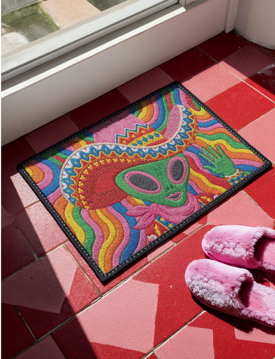 A rectangular doormat featuring a vibrant 2D flat print designed to look like textured yarn embroidery. The central graphic showcases a bright green alien with large black eyes, wearing a pink neck bandana and a highly decorated red, pink, and blue sombrero cowboy hat. The alien has one green hand raised in a friendly wave. The background is a psychedelic pattern of wavy, flowing rainbow stripes in yellow, orange, red, pink, blue, and green. The mat has a sleek black outer edge trim.