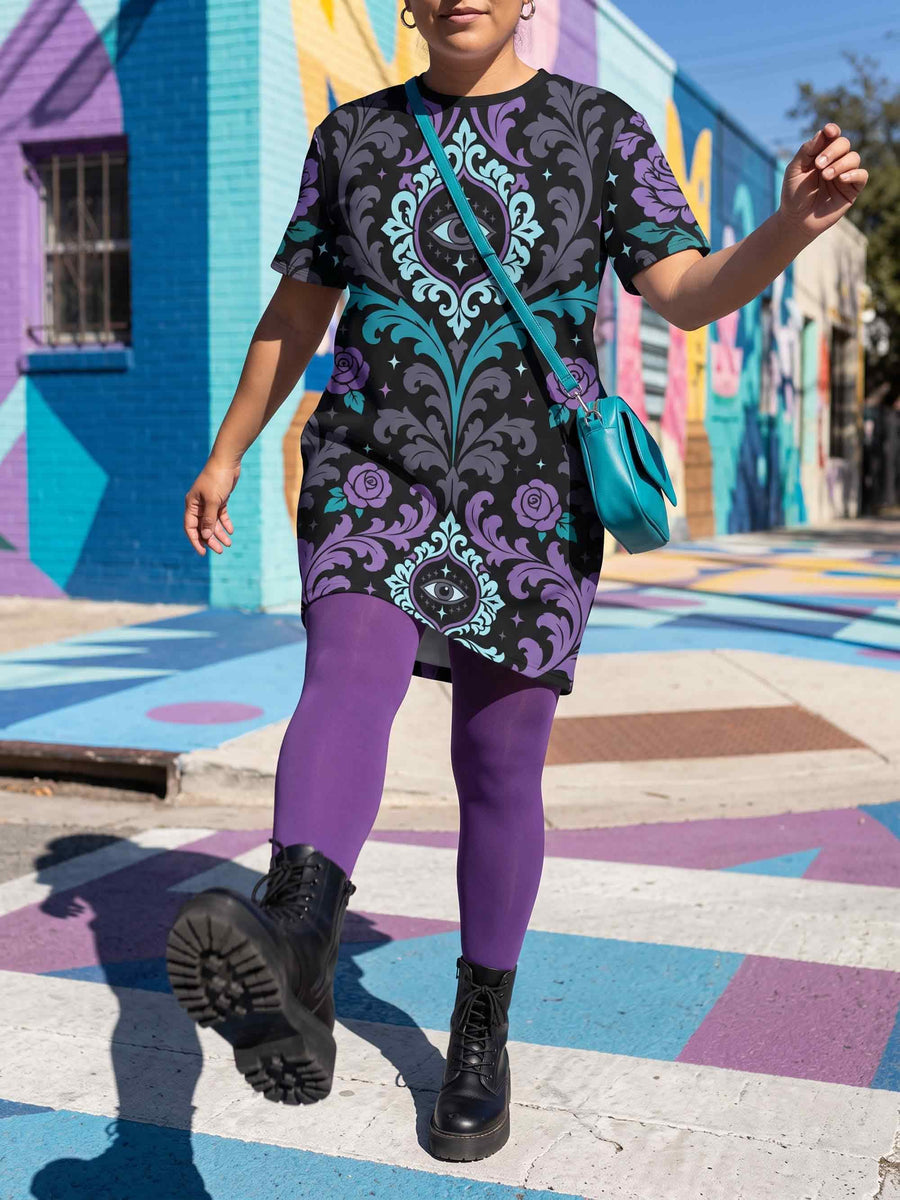 Cotton t-shirt dressA short-sleeve structured t-shirt dress with a black background. The dress features a bold, vertically stacked repeating graphic print. The design includes intricate damask patterns in teal and grey, purple roses with teal leaves, and a central evil eye motif set inside an ornate frame with small four-point stars. The heavyweight fabric holds a clean, straight cut above the knee with zero drape, maintaining a sculptural, slightly boxy silhouette.