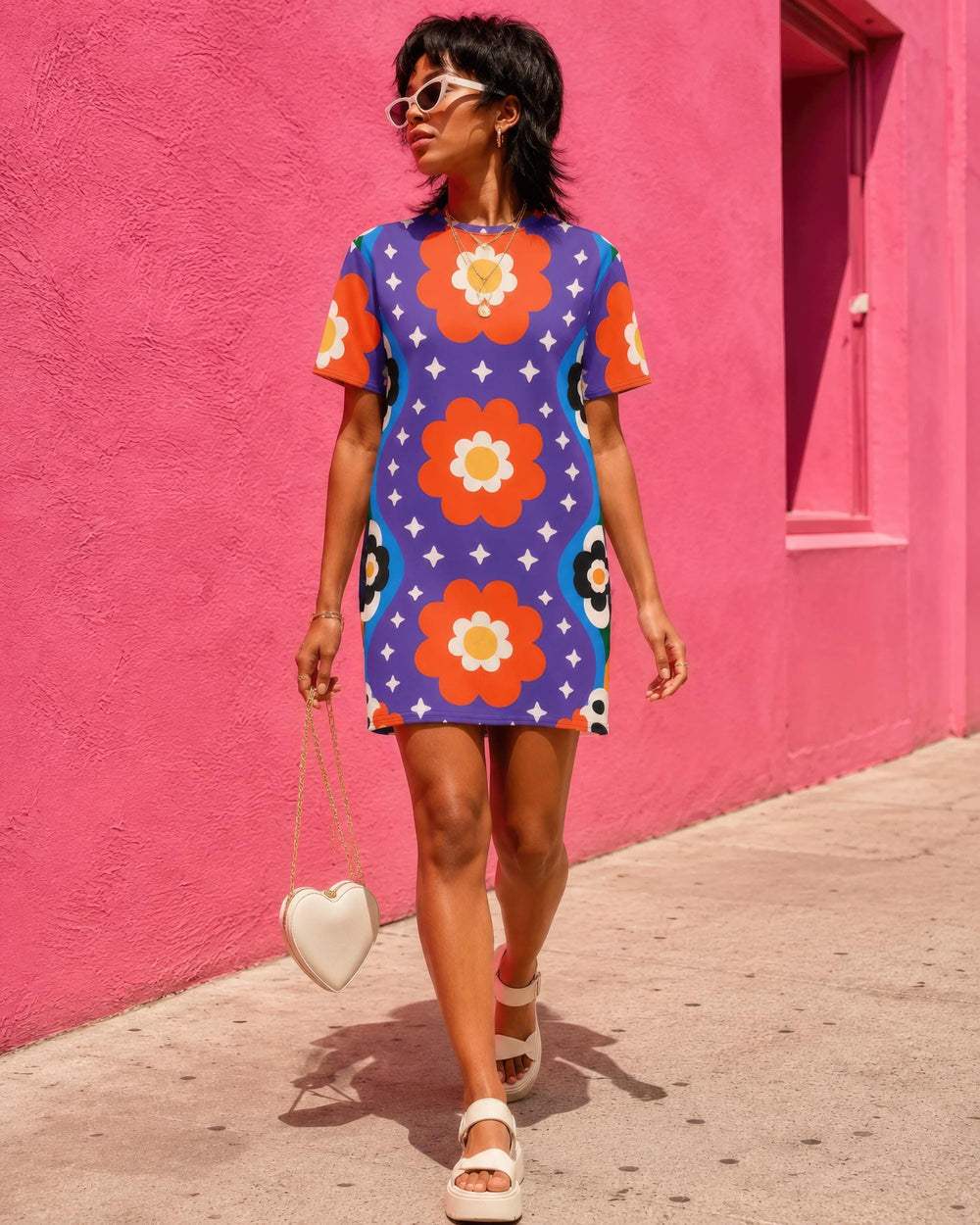 Short sleeve crew neck t-shirt dress in a relaxed, oversized straight cut, made of 100% polyester with a soft matte finish. An edge-to-edge all-over print on a medium purple background shows vertically stacked daisies with orange petals and yellow centers, scattered white four-point stars, and blue and green wavy side borders filled with small black and white daisies. Retro 70s floral, groovy maximalist alt fashion.