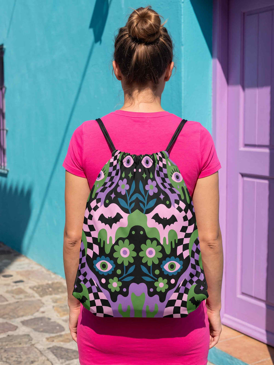 Drawstring backpack measuring 15 by 17 inches, made of 100% polyester with narrow shoulder straps and a drawstring closure. A symmetrical pastel goth print of bats, stylized eyes, flowers, and stars in a soft pastel palette sits on a black base, framed by purple and green checkerboard bands. Creepy cute, whimsigoth, kawaii goth style bag made for the gym, concerts, festivals, travel, school, and everyday carry.