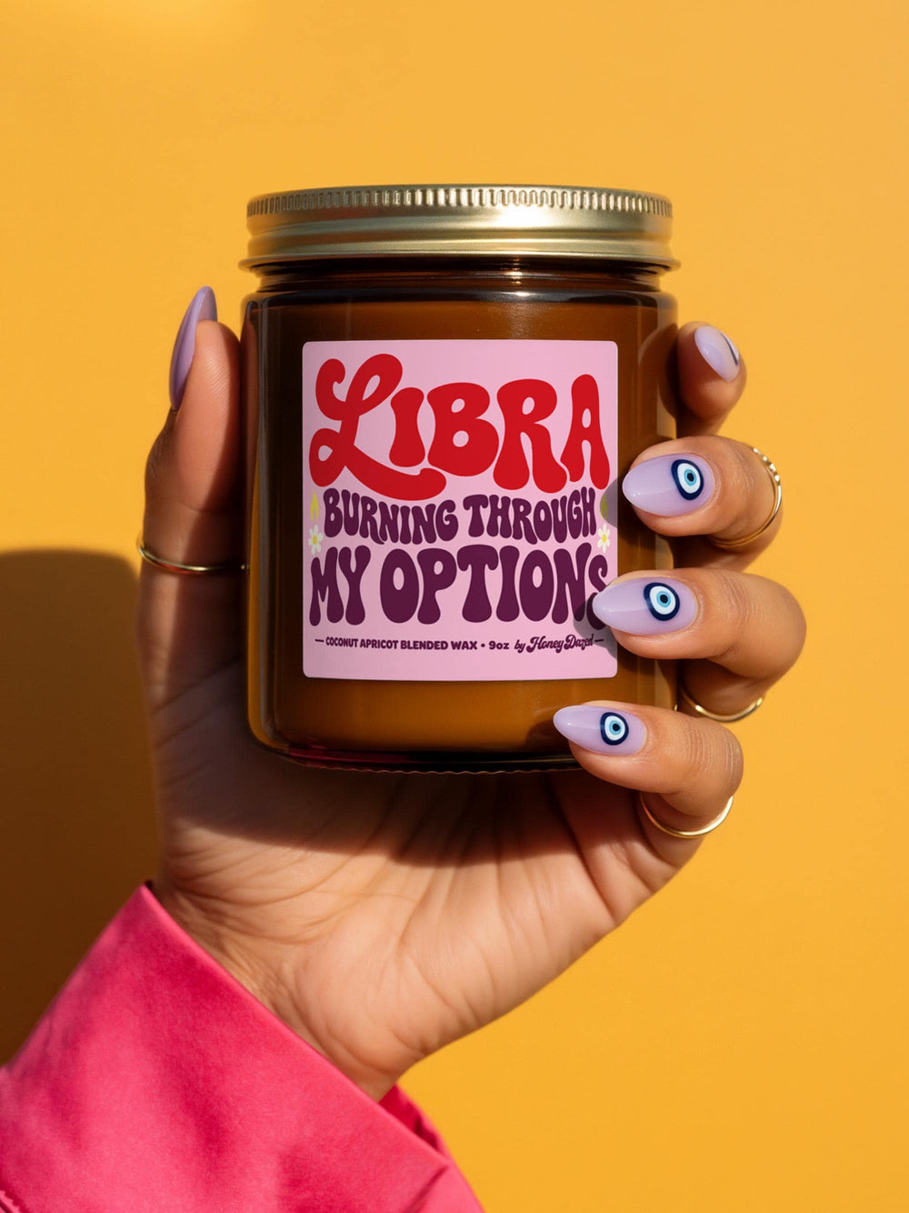 Funny Libra zodiac candle in a 9 oz amber glass jar with a brushed gold screw lid. Bubblegum pink label with Libra in big groovy 70s retro red lettering, the phrase BURNING THROUGH MY OPTIONS in wavy purple and red typography, and tiny yellow and green daisies, coconut apricot wax 9oz by Honey Dazed at the bottom. Whimsical dopamine decor astrology candle made for a nightstand, desk, or bath shelf, a Libra birthday gift.