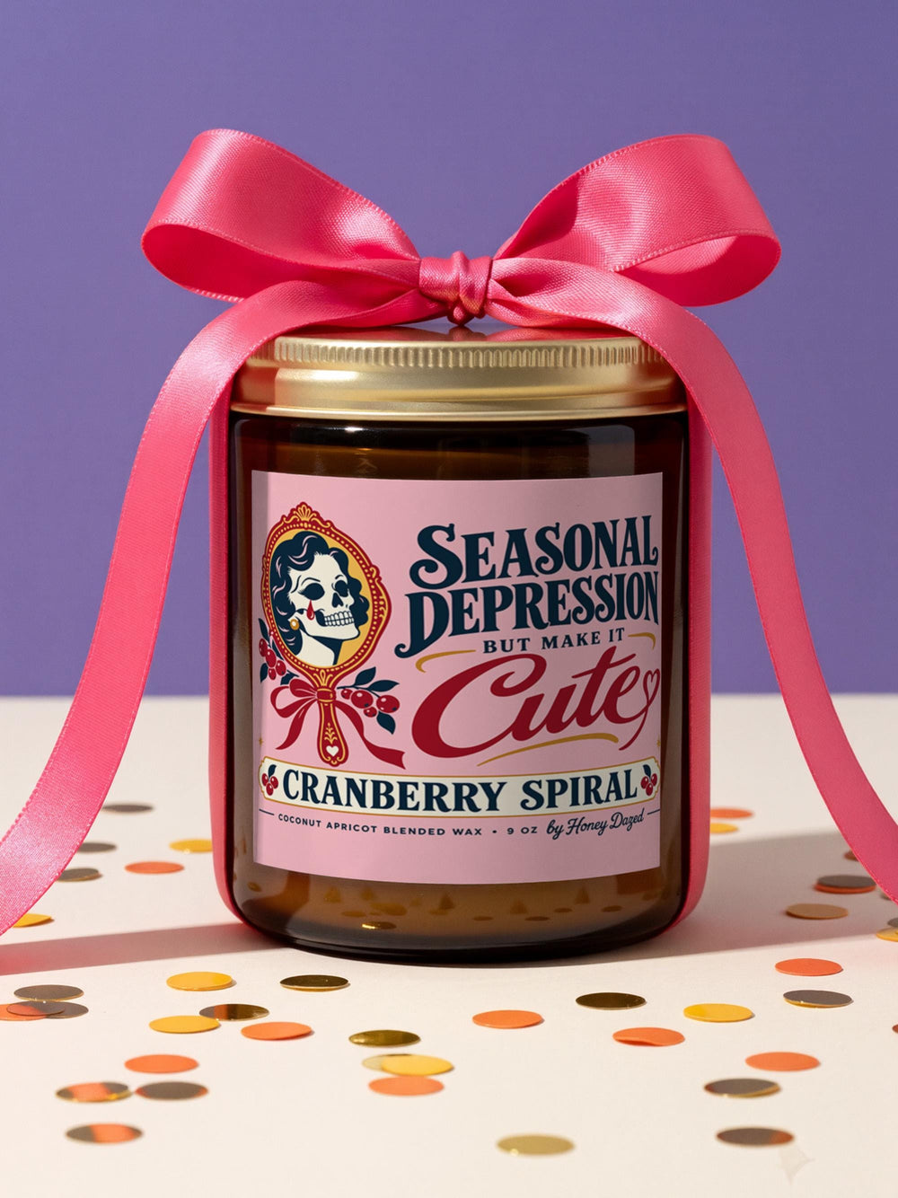 Funny Seasonal Depression candle in a 9 oz amber glass jar with a brushed gold screw lid. Pale pink label with an ornate crimson and gold hand mirror framing a retro pinup skull shedding a single tear, cranberry branches, and a red ribbon bow, with navy and cherry red type reading Seasonal Depression But Make It Cute and Cranberry Spiral. Dark coquette, whimsigoth, tattoo flash, dark humor decor made for a nightstand, vanity, or windowsill.