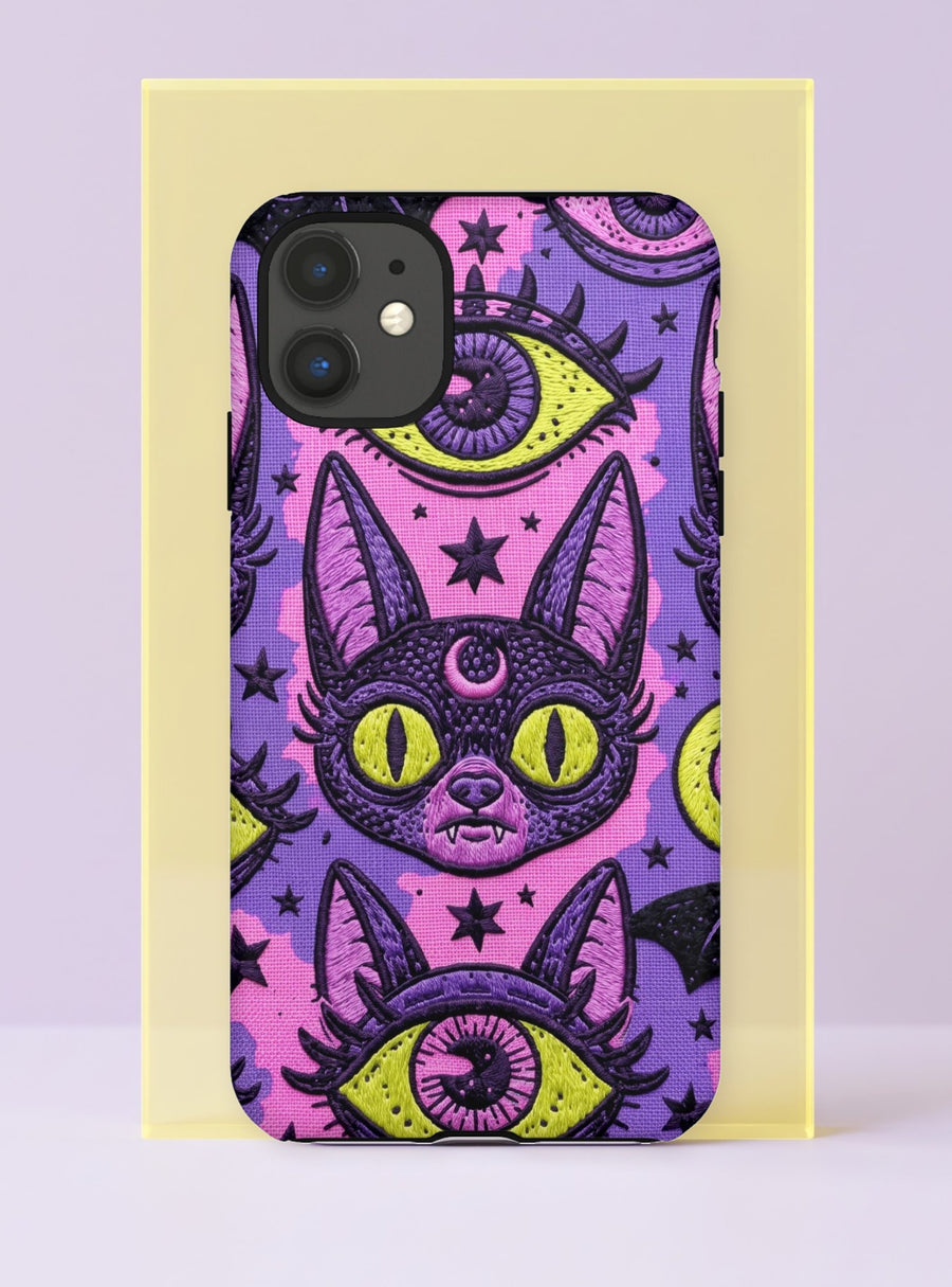 Dual layer matte phone case with a 2D print that mimics raised embroidery. Dark purple vampire bat cat heads with tall pointed ears, glowing chartreuse eyes, tiny fangs, and a pink crescent moon on the forehead repeat across a mottled purple and hot pink background, scattered with large chartreuse evil eyes with purple irises and small dark stars. Smooth printed surface with no raised threads, open ports, and camera cutout. Pastel goth, whimsigoth, creepy cute phone accessory.