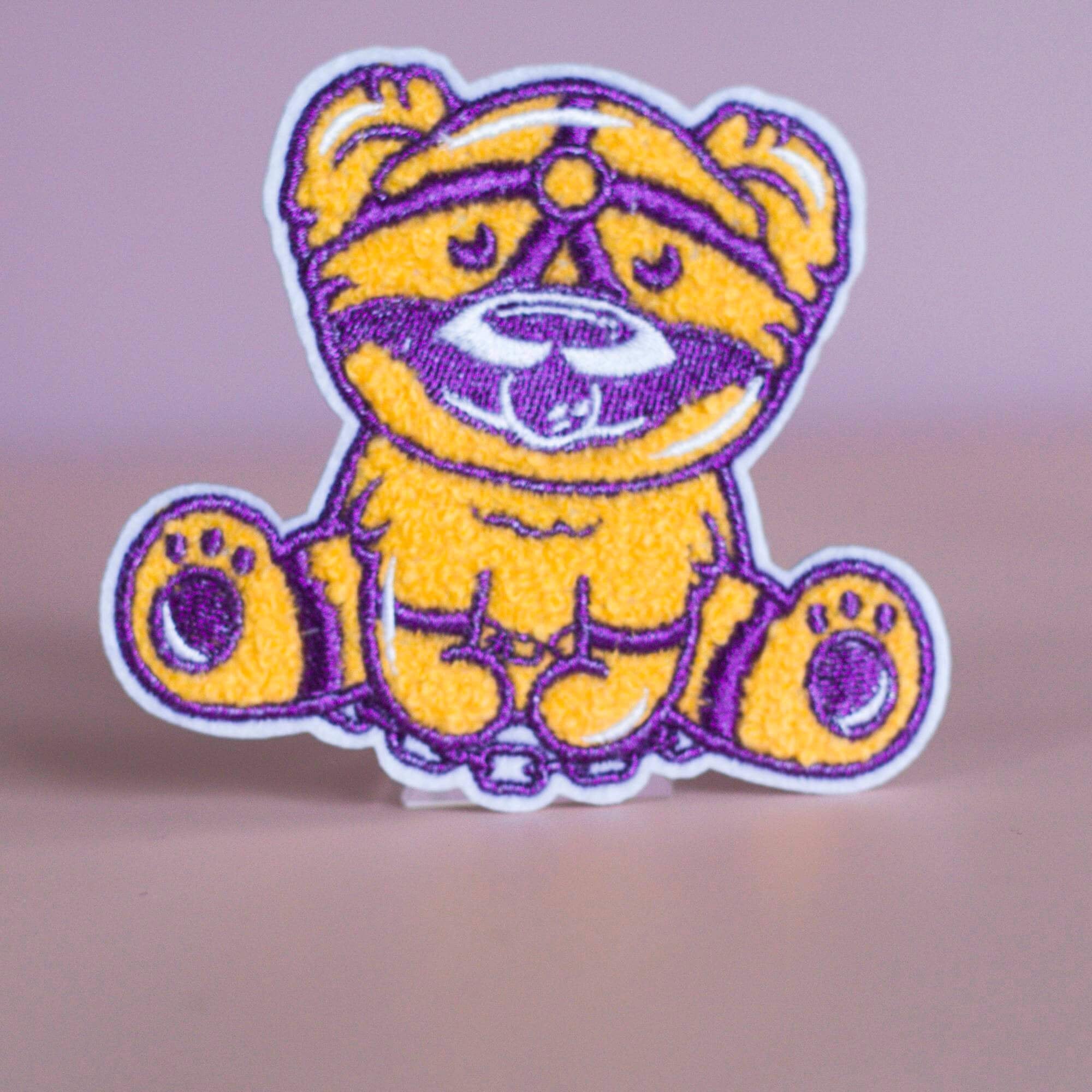 Patch featuring a cute bear in yellow and purple with a playful expression, wearing a BDSM harness and a ball gag. The design is fun and whimsical, combining bold colors with a cheeky style.